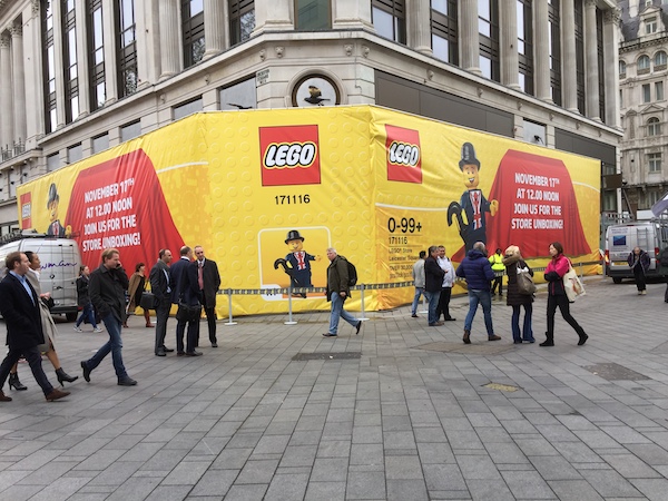A photo of a building hoarding in a city square, with a large Lego logo and promotional banners.