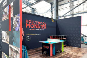 A Hollywood Monster exhibition booth with a table tennis table, a branded black wall, and a side panel with fragmented images.