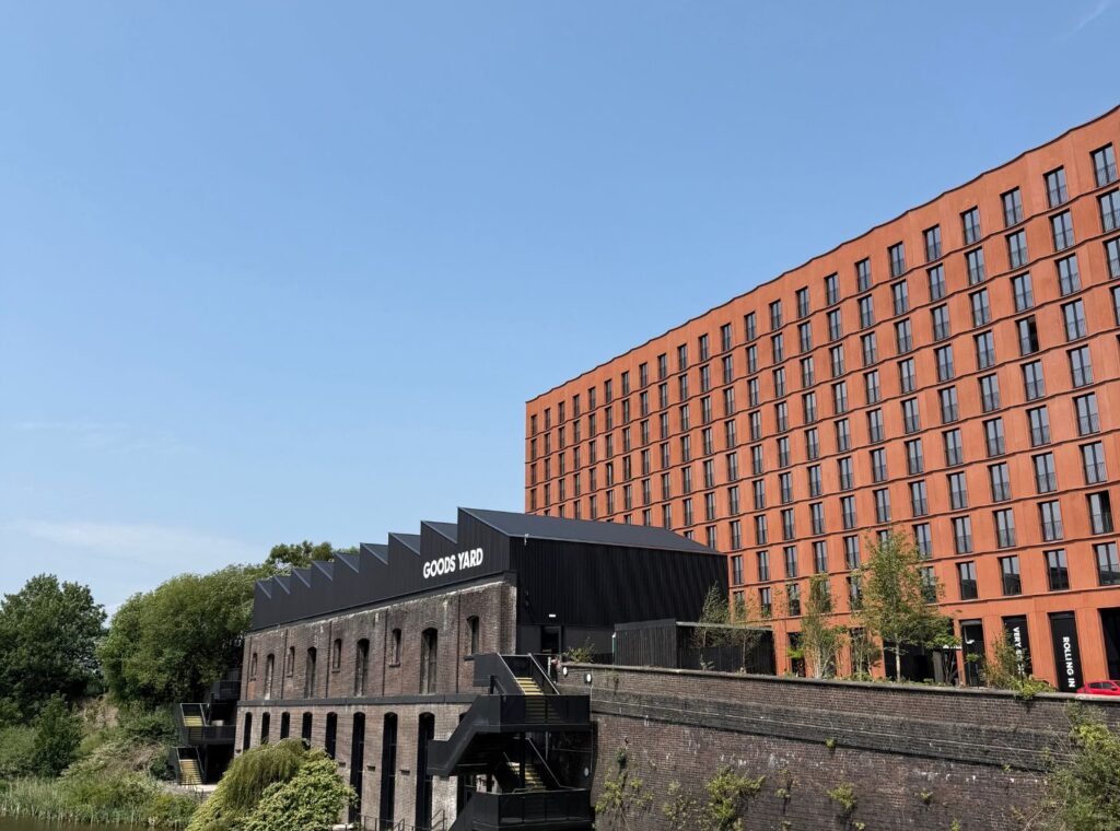 A modern, tall orange building with many windows stands behind a lower, older brick building with "GOODS YARD" signage