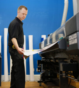 A man in a black shirt inspects a printout from a large digital printer, standing between two similar machines in a room with a vibrant blue wall.