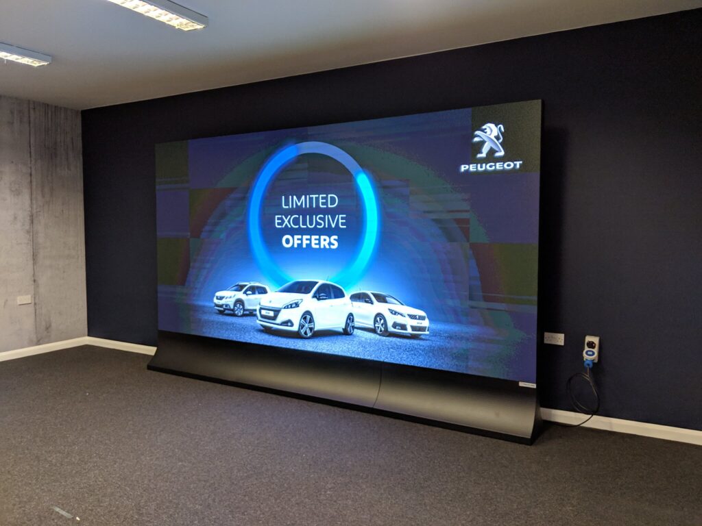 A large, curved digital display screen is showing automotive signage for Peugeot, with the brand's logo and "LIMITED EXCLUSIVE OFFERS" text.