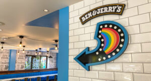 A Ben & Jerry's ice cream shop features a white subway tile wall with a prominent "BEN & JERRY'S" sign and a large blue arrow sign