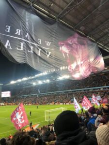 Large format print banner with a wolf head and text hanging in a packed football stadium, with smaller flags in the crowd.