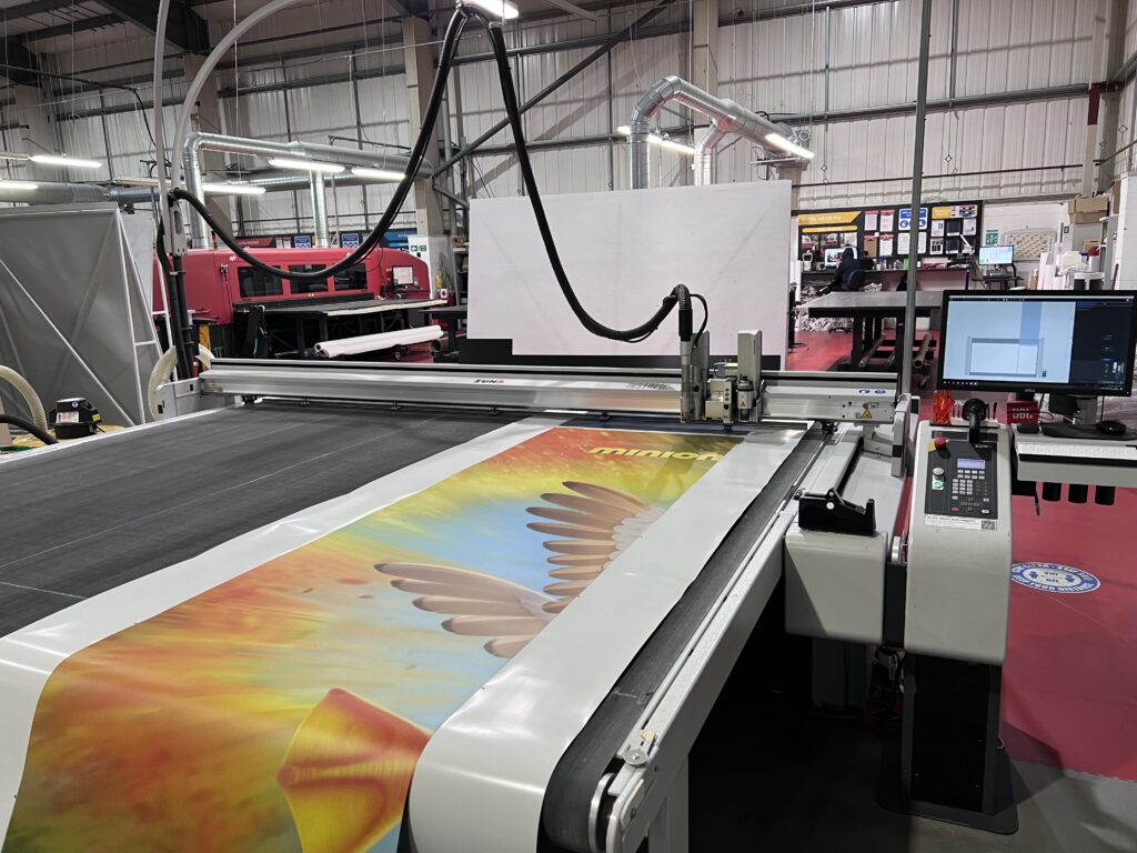A large, wide-format digital printer is printing a colorful graphic of a phoenix or eagle on a long roll of white material.