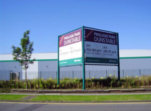A large double-sided sign for "PROLOGIS PARK DUNSTABLE" stands on a grassy median beside a road. The sign advertises two units, DC1 and DC2, with their respective square footage. In the background, there's a large white industrial building with a green stripe, and a young tree is planted to the left of the sign. The sky is clear and blue.