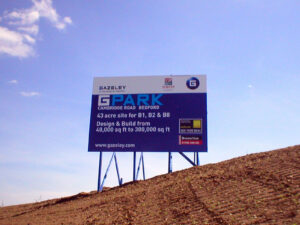 A large blue and white billboard advertising "G PARK Cambridge Road Bedford" on a brown dirt mound against a blue sky.