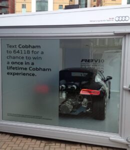 A small, glass-enclosed outdoor display case features automotive signage for an Audi R8, with the back half of the car visible and a text-in competition.