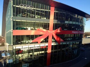 A modern, multi-story glass building with a car dealership showroom is decorated for Christmas with a giant red ribbon graphic and a bow wrapped around the facade.
