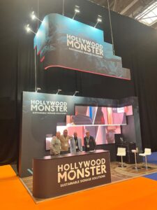 A display booth for "Hollywood Monster" at a trade show, featuring four men standing behind a branded counter. The booth has large signage that reads "HOLLYWOOD MONSTER SUSTAINABLE SIGNAGE SOLUTIONS."