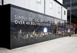 A temporary hoarding boards at a construction site, featuring a large black graphic. The graphic shows a stylised city skyline and a woman with long red hair. The text reads 'SIMPLY DELICIOUS' and 'OVER 40 BOUTIQUE EATERIES'.