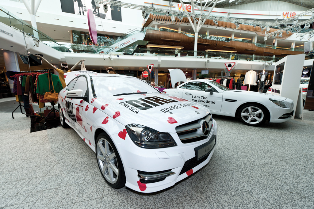Two white Mercedes-Benz cars are on display in a shopping centre. The car in the foreground has custom vinyl graphics with red hearts and text