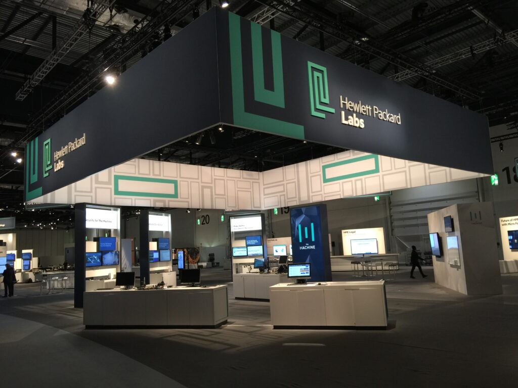 An exhibition space for Hewlett Packard Labs at ExCeL London. A large, dark blue hanging sign with green lines and the company name is suspended over the main display area. The space is filled with multiple white display stands featuring monitors and technology.