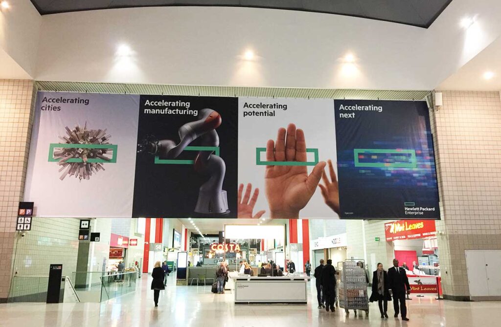 A large, multi-panel banner for Hewlett Packard Enterprise hangs in a shopping mall or convention centre. The banner is divided into four sections, each with a different graphic and a title: 'Accelerating cities,' 'Accelerating manufacturing,' 'Accelerating potential,' and 'Accelerating next.' The banner features abstract images of a city, a robotic arm, and hands. Below the banner, people are visible walking through a brightly lit concourse with shops like Costa and M&S.