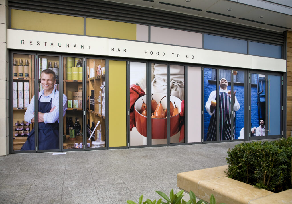 The storefront of a restaurant or bar is covered with a series of vertical window graphics. The graphics feature close-up photographs of smiling chefs and food items, with the words 'RESTAURANT', 'BAR', and 'FOOD TO GO' written above the doors.
