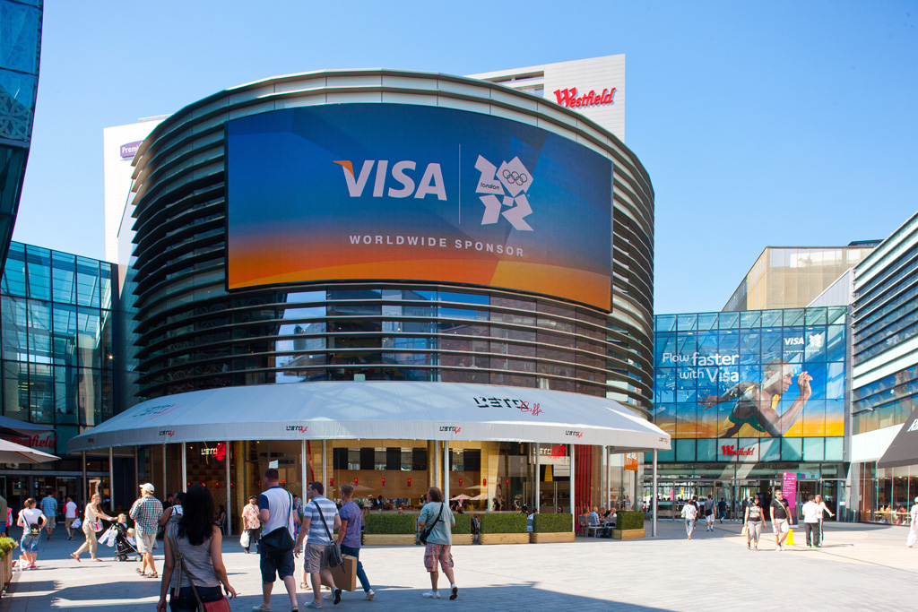 A large, curved building at a Westfield shopping centre features a huge LED screen displaying a Visa athletic signage advertisement for the London 2012 Olympics.