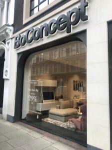 The storefront of a BoConcept furniture store, with a modern living room display and a sign for a sale of up to 20% off.