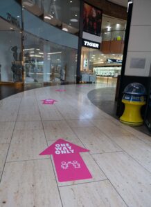 Bright pink "One Way Only" arrows on a tiled floor in a shopping mall to guide foot traffic during the COVID-19 pandemic.