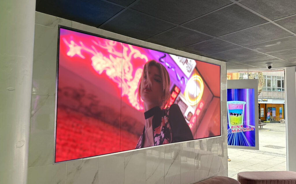 A large digital video wall and a smaller vertical digital screen displaying advertisements in an indoor space.