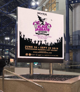 A large LED screen in a public space displaying a poster for the "Black Sabbath - 50 Years" exhibition at the Birmingham Museum & Art Gallery.