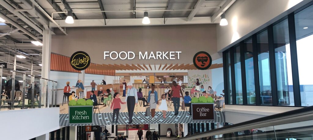 A large, brightly coloured mural inside a Sainsbury's store, depicting a lively "Food Market" with stylised illustrations of people and various food stalls.