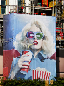 large building wraps that features a stylised image of a blonde woman wearing large, mismatched blue and green 3D glasses. She holds a striped box of popcorn in one hand and sips from a soda cup with a straw in the other. The banner is wrapped around scaffolding at a construction site in a city.