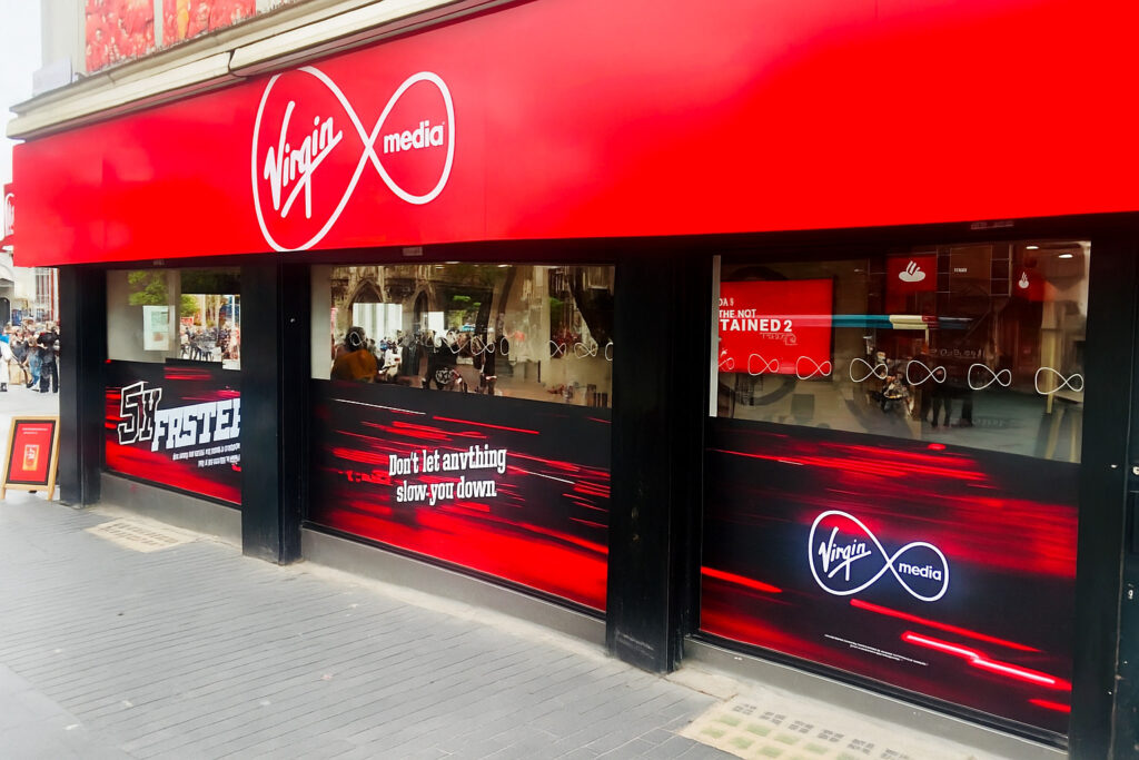 The exterior of a Virgin Media store, featuring a bright red sign with the company's logo. The lower half of the storefront windows is covered with black and red window graphics showing abstract red lines and the slogan 'Don't let anything slow you down'. The brand logo is repeated on the graphics.