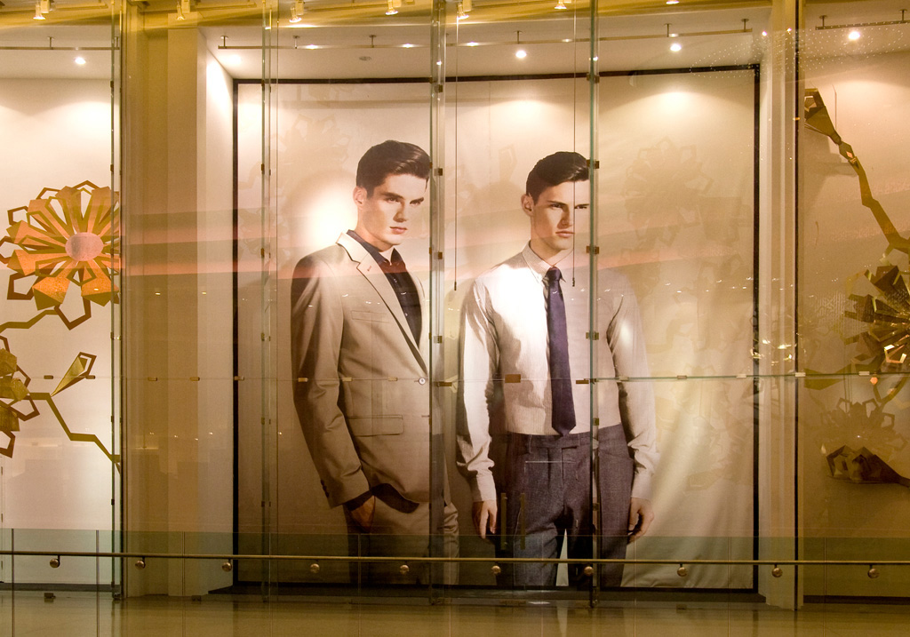 Two large-scale, full-length portraits of male models are displayed as temporary window graphics in a mall. The models are dressed in formal wear, one in a tan suit and the other in a light-coloured shirt and tie. The graphics are reflected in the glass, and other store displays are visible to the sides.