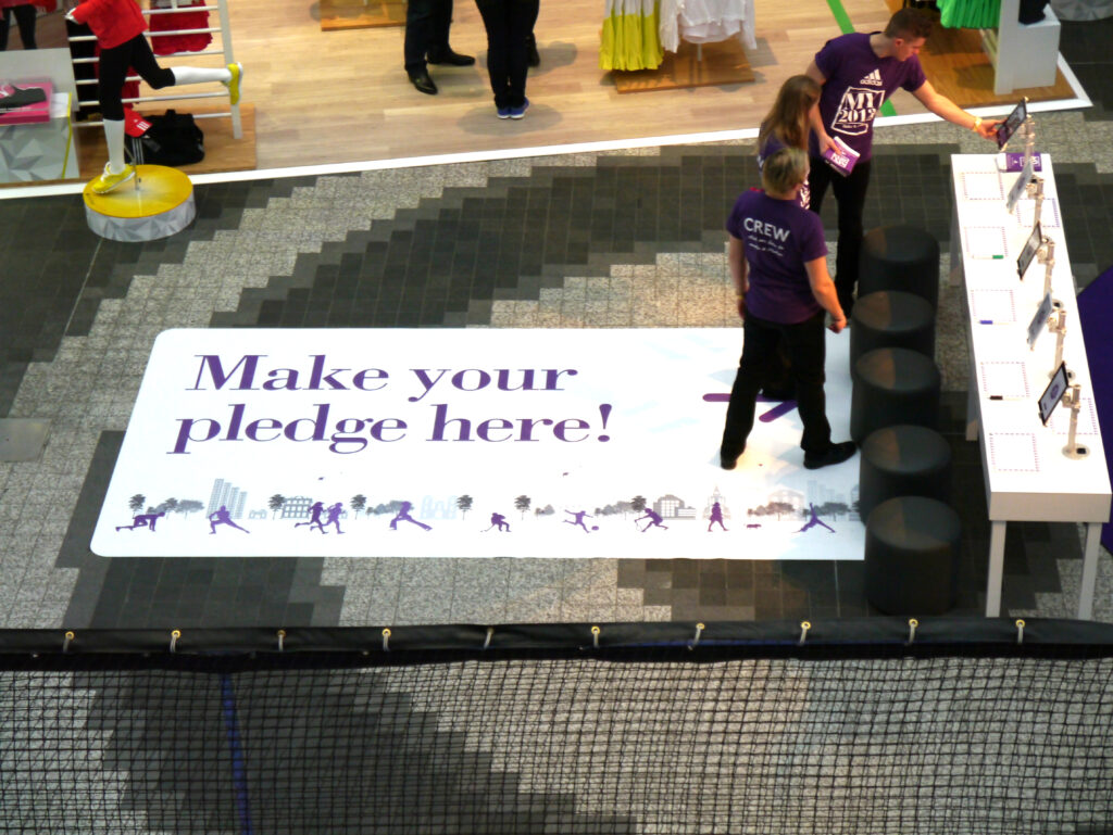 A high-angle view of a promotional stand in a shopping centre with a floor graphic that reads "Make your pledge here!", with staff in purple shirts helping customers.