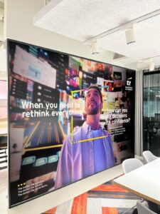 A very large video wall or display screen in a modern office, featuring the EY logo and a striking advertisement. The screen shows a man looking upwards against a vibrant, abstract city background, with the text "When you need to rethink everything" and "how can you make decisions with confidence?". This serves as dynamic corporate signage and internal branding.