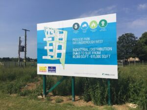A large outdoor billboard, acting as industrial & logistics signage, advertises Prologis Park Wellingborough West. The sign features a site plan, key benefits (value, size, labour, quality), and promotes "INDUSTRIAL/DISTRIBUTION BUILD TO SUIT FROM 80,000 SQ FT – 615,000 SQ FT." The board is situated in a grassy field under a bright blue sky, with a utility pole to the left.