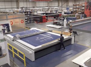 A wide shot of a factory floor shows two workers operating a large flatbed cutting machine that is processing a blue material with white graphics. One person is positioned at the side of the machine, holding a long cylindrical tool, while another person stands near a computer console at the end of the machine. In the background, there are various other industrial machines, rolls of material, and storage racks, indicating a large-scale production facility.