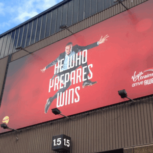 Virgin Trains billboard on a corrugated building showing an image of a man in a suit leaping mid-air with the slogan 'HE WHO PREPARES WINS' on a red background.