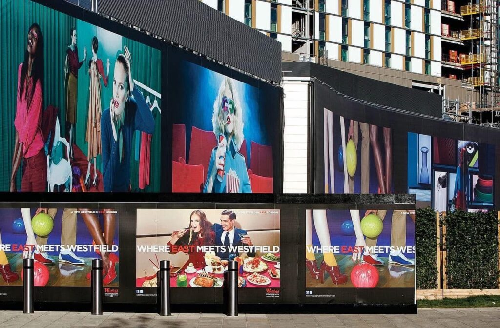Large, colourful fashion and lifestyle advertising panels on construction hoarding for Westfield Stratford City in London.