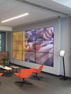 Indoor video wall comprised of 9 screen, with a video of a chef cutting peppers, in front of the screen is two orange chairs