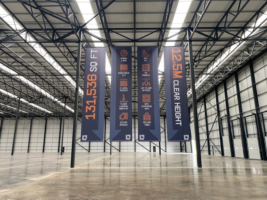 Warehouse operational signage, 4 large banners in a warehouse, one that reads 131,536 sq ft, the banner next to that pewr 1 mva, euro dock, 2 level access, floor loading 50kn/m2, 19 hgv spaces, the banner next to that reads epc a+, pv panels, 1st floor offices, site coverage 45%, secure yard, the banner on the far right reads 12.5m clear height.