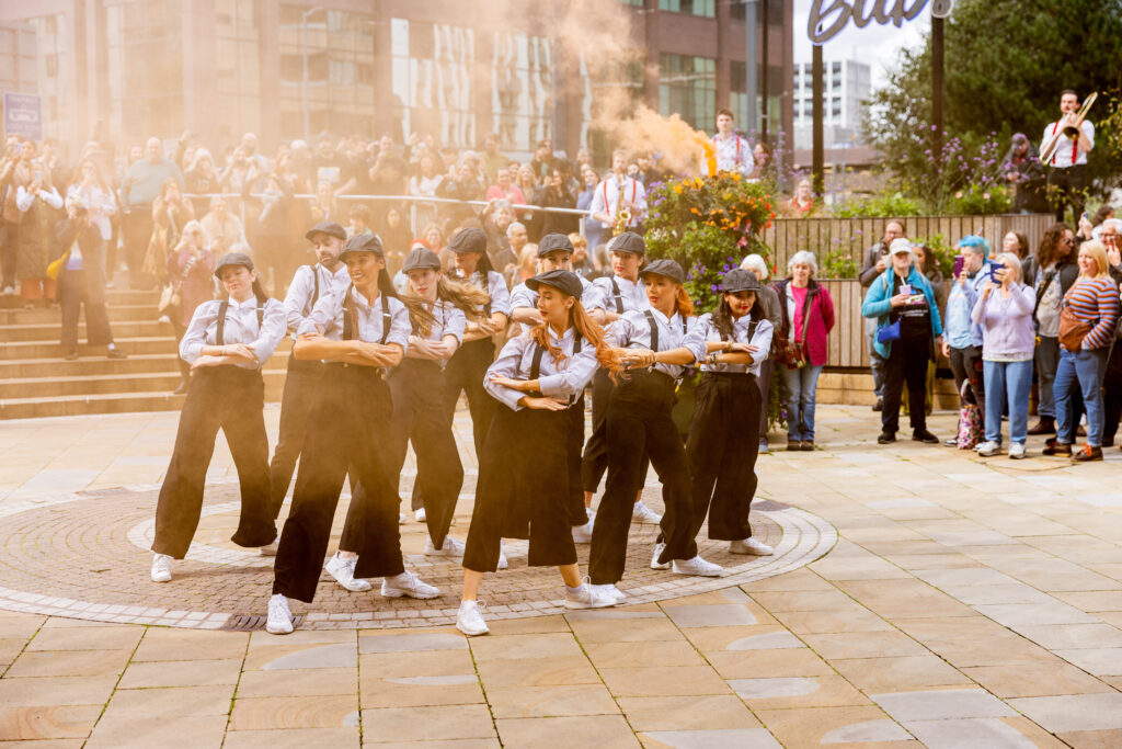 A group of dancers dressed in a suit and a flat cap with a group of onlookers behind them at the international day of Birmingham